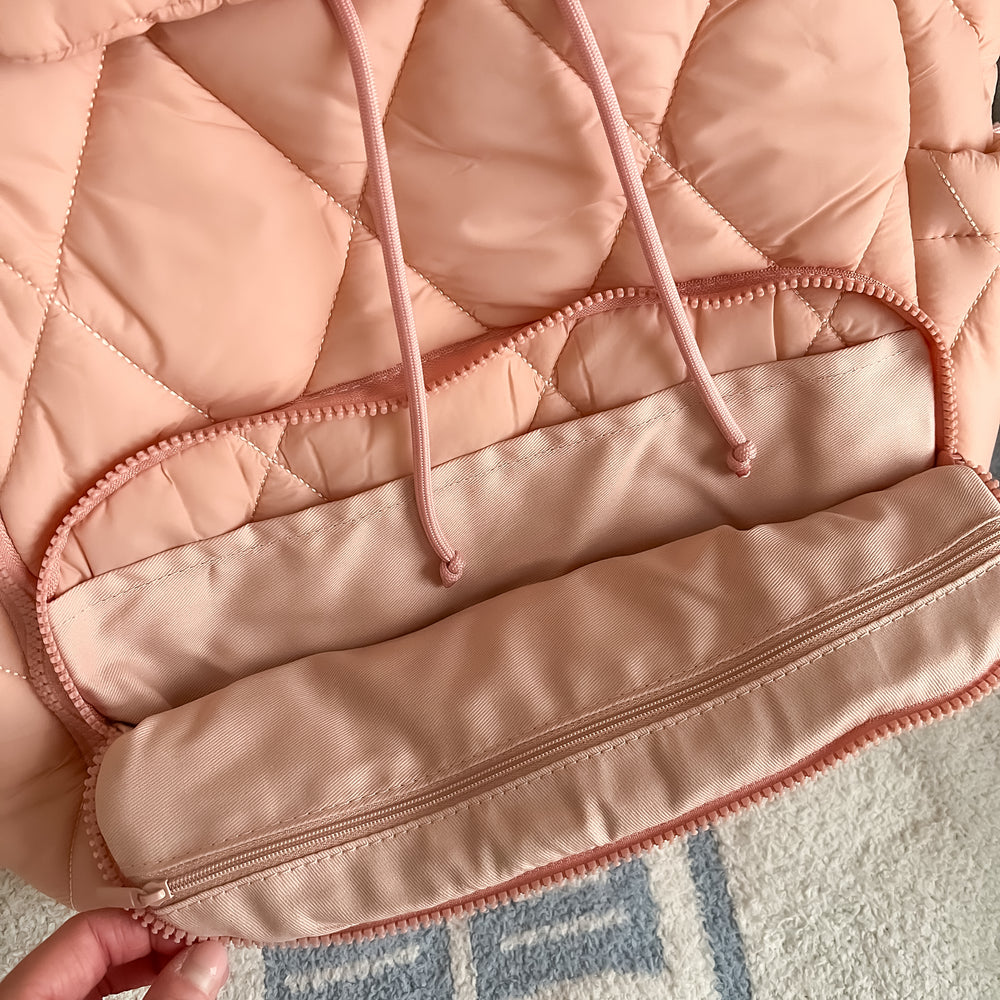 Close-up of a pink blush bookish backpack quilted jacket with a zipper, held by a hand on a textured surface.